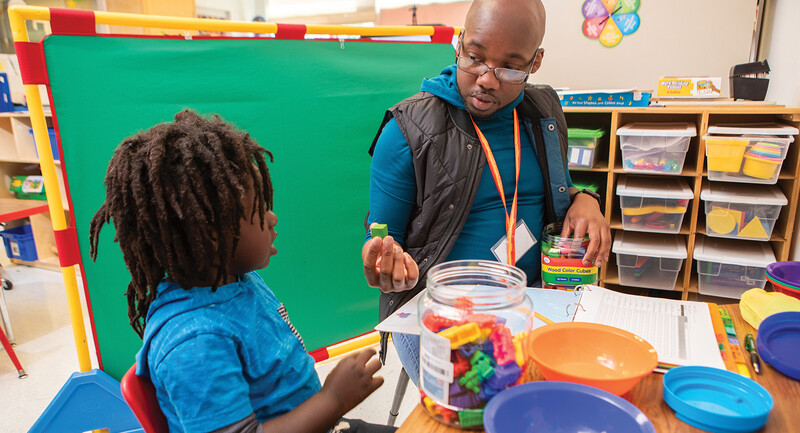 A teacher and student sitting at a desk in a colorful classroom working with small wooden cubes to improve mathematical understanding 