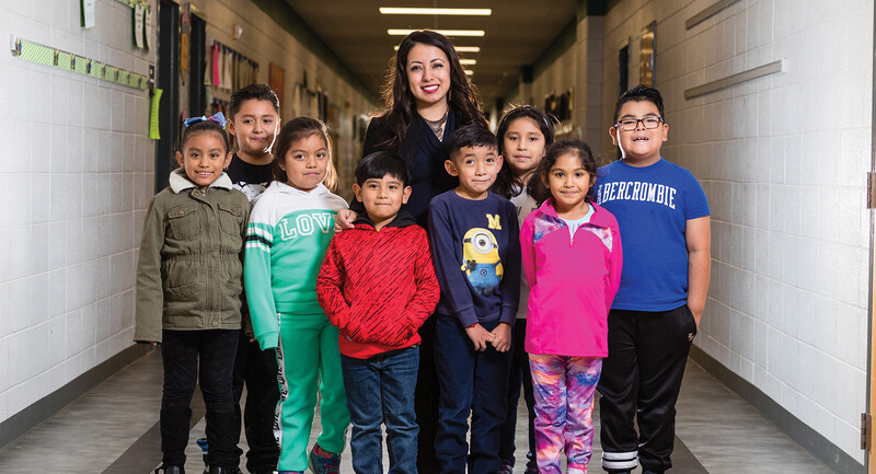 A smiling teacher stands in a hallway surrounded by eight happy students in a group